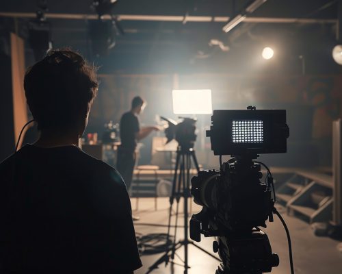 A man stands in front of a camera with a light shining on him A man stands in front of a camera with a light shining on him. He is wearing a black shirt and he is a director or a cameraman. The scene is set in a dark room with a spotlight on the man